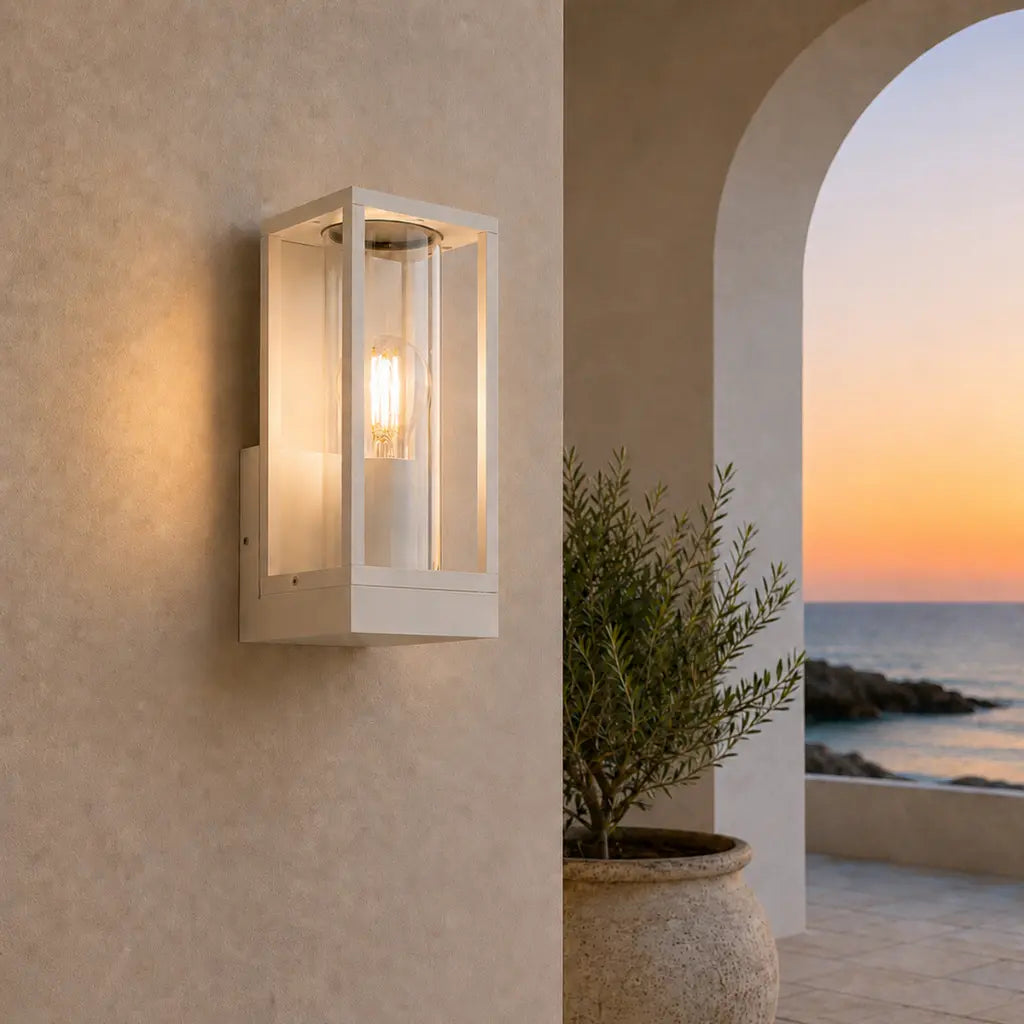 A modern rectangular outdoor wall sconce with a white frame and warm glowing light, mounted on a beige stucco wall next to a potted plant and an arched view of the ocean at sunset.