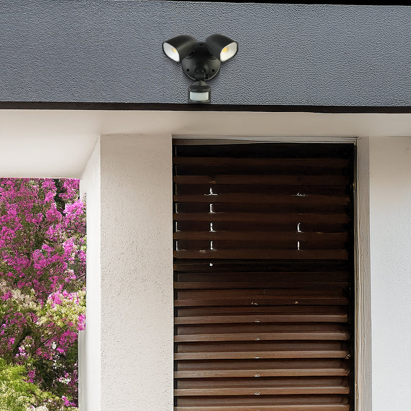 Outdoor security light fixture on a house exterior with flowers in the background