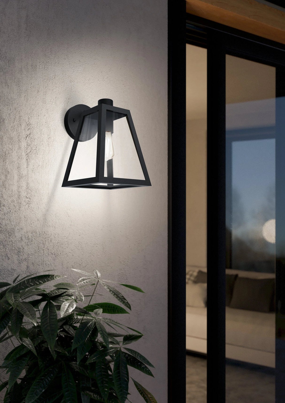 Black outdoor Sleek And Modern Wall Sconce Eglo MIRANDOLA on a textured wall with a plant in the foreground.