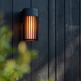 Wall-mounted outdoor light fixture on a dark wooden wall with blurred greenery in the foreground.