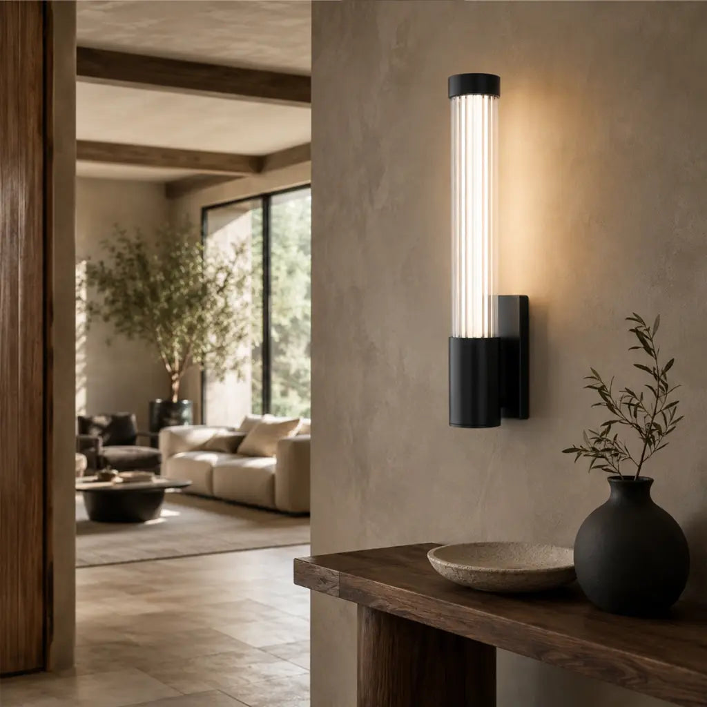 A modern wall-mounted light fixture with a cylindrical glass shade and black metal base, glowing warm white on a beige wall in a styled interior hallway.
