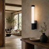 A modern wall-mounted light fixture with a cylindrical glass shade and black metal base, glowing warm white on a beige wall in a styled interior hallway.