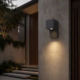 A modern dark gray rectangular outdoor wall sconce mounted on a textured concrete wall, casting a warm light downward in front of a contemporary house entrance at dusk.