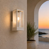 A modern rectangular outdoor wall sconce with a white frame and warm glowing light, mounted on a beige stucco wall next to a potted plant and an arched view of the ocean at sunset.