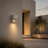 A modern square outdoor wall light fixture mounted on a textured beige stucco wall, casting a warm yellow light downward. The scene is set in a minimalist architectural courtyard with a tree in a planter and soft evening sky in the background.