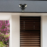 Outdoor security light fixture on a house exterior with flowers in the background