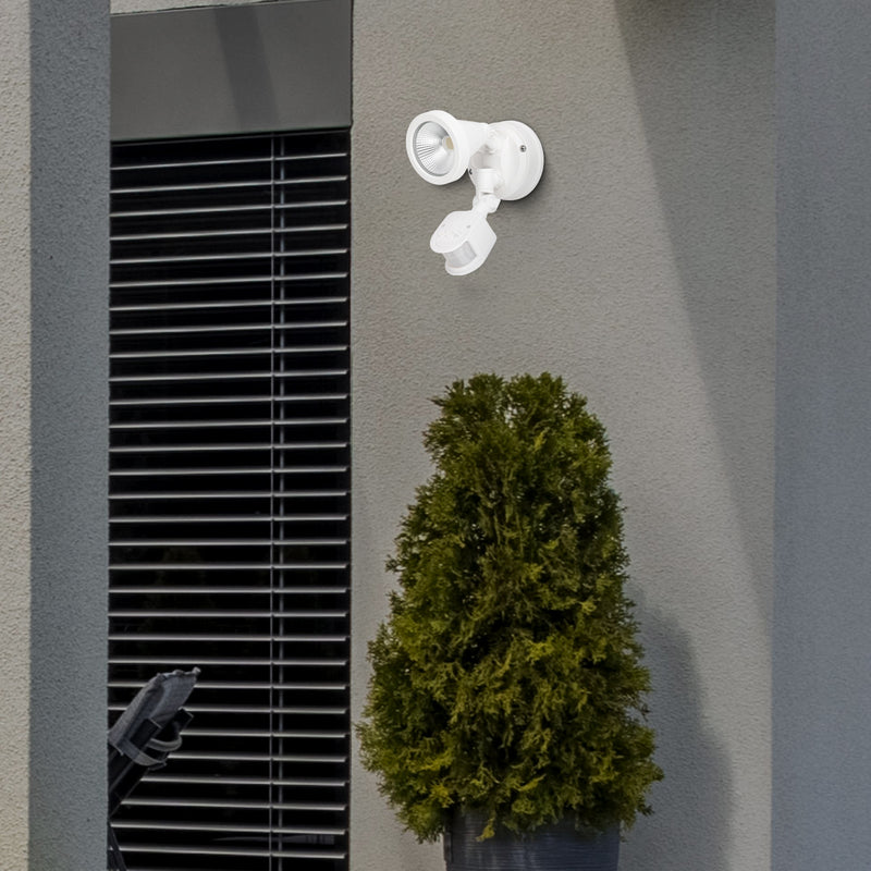 White outdoor security camera mounted on a gray wall next to a green bush.